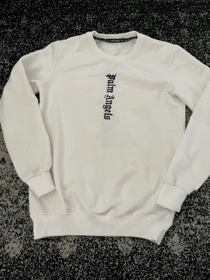 Vit sweatshirt från Palm Angels - Säljer en clean vit sweatshirt från Palm Angels med svart logotyp i gotisk stil både fram och bak. Tröjan har rund hals, ribbade muddar och är tillverkad i mjukt bomullsmaterial. Perfekt för dig som gillar streetwear och vill ha en stilren look.