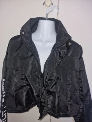 Snygg svart vindjacka - Svart croppad vindjacka från PrettyLittleThing med hög krage, dragkedja. Jackan har flera stora fickor framtill och coola detaljer. Perfekt för dig som gillar streetstyle och vill sticka ut.