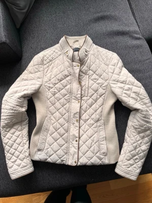 Beige quiltad jacka med ståkrage - Snygg beige quiltad jacka med ståkrage och guldfärgade knappar. Jackan har långärmad design, ribbade sidopaneler och två fickor framtill. Perfekt för dig som gillar stilrena detaljer och en modern look.