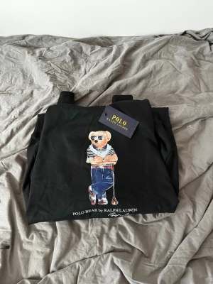 Svart hoodie Polo Ralph Lauren L - Svart hoodie från Polo Ralph Lauren med den ikoniska Polo Bear-trycket framtill. Klassisk huva med dragsko och ribbade muddar. Tillverkad i mjukt bomullsmaterial för skön känsla. Perfekt för dig som vill ha en stilren och trendig look.