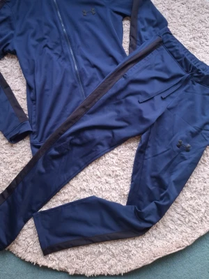 Marinblå träningsset från Under Armour - Säljer ett marinblått träningsset från Under Armour med svarta detaljer längs ärmar och ben. Setet består av en ziptröja och matchande byxor i mjukt och stretchigt syntetmaterial, perfekt för träning eller uppvärmning.