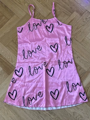 Rosa nattlinne med hjärtan och text - Sött rosa nattlinne i satinlook med tunna axelband. Tryckt med svarta hjärtan och ordet 'love' över hela plagget. Kort modell med avslappnad passform, perfekt för mysiga kvällar. Materialet känns mjukt och glansigt mot huden.