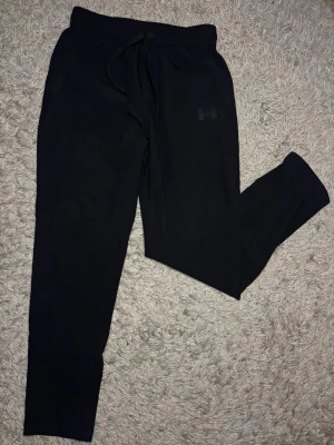 Svarta mjukisbyxor från Under Armour - Säljer ett par svarta mjukisbyxor från Under Armour med dragsko i midjan och elastisk resår. Byxorna har en diskret logga på låret och är tillverkade i ett mjukt och bekvämt material. Perfekta för chill eller träning. Storlek M men passar som S