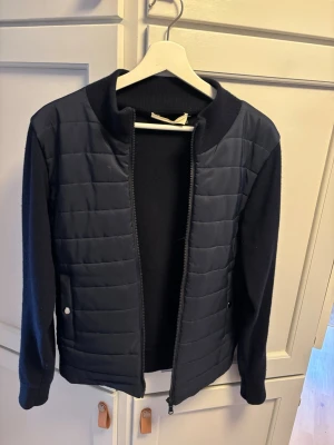 Mörkblå cardigan - Snygg mörkblå cardiganjacka med quiltad framsida och mjuka stickade ärmar. Jackan har dragkedja framtill, ribbad krage och två fickor med knapp. Perfekt för lager på lager och en clean look. Den är storlek S men passar xs då den är lite liten o storleken. Priset är inte hugget i sten!