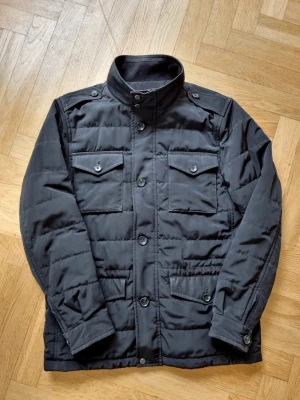 Svart pufferjacka från Riley - Snygg svart pufferjacka från Riley med hög krage, fyra framfickor med lock och knappar samt axelklaffar. Jackan har quiltad design och stängs med både dragkedja och knappar. Perfekt för kalla vinterdagar och ger en stilren look.