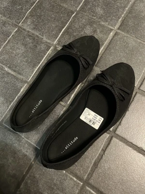 Svarta ballerinaskor med rosett - Helt nya stilrena svarta ballerinaskor från Attitude med söt rosett framtill. Använda en gång❣️Skorna har rund tå och är tillverkade i ett tygmaterial med diskret mönster. Perfekta för en enkel och klassisk look. Säljer pga att de var för små❣️