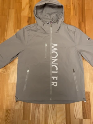 Grå vindjacka från Moncler - Snygg grå vindjacka från Moncler med stor vit logga längs dragkedjan framtill. Jackan har huva, tre framfickor med dragkedja och Moncler-märke på ärmen. Tillverkad i lätt, vattenavvisande material och har justerbar nederkant.