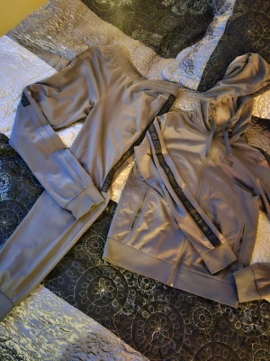 Grått set från EA7 Armani - Snyggt grått set från EA7 Emporio Armani med svarta logotyper och detaljer längs sidorna. Byxorna har resår i midjan och muddar vid bensluten för en skön passform. Matchande hoodie medföljer, båda i mjukt och glansigt material. Små hål på byxorna som sista två bilderna visar som knappt märks.