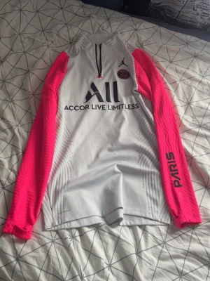 PSG x Jordan träningströja vit/rosa - Snygg långärmad träningströja från PSG x Jordan i vitt med neonrosa ärmar och svarta detaljer. Halvlång dragkedja framtill, Paris-tryck på ena ärmen och klubbmärke på bröstet. Tillverkad i ett lätt och ventilerande material, perfekt för träning.