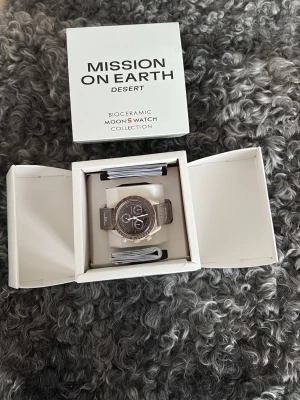 Swatch x Omega Desert - Säljer en Swatch x Omega MoonSwatch Mission on Earth Desert med beige och bruna toner. Klockan har biokeramisk boett, tydlig urtavla med kronograf och matchande textilarmband. Kommer i originalbox med papper. Perfekt för dig som gillar unik design.