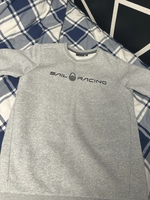Grå sweatshirt från Sail Racing, 176 men passar S och XS - Snygg grå sweatshirt från Sail Racing med svart logotyp och text på bröstet. Tröjan har rund halsringning, långa ärmar och är tillverkad i mjukt bomullsmaterial. Perfekt för en avslappnad och sportig stil.