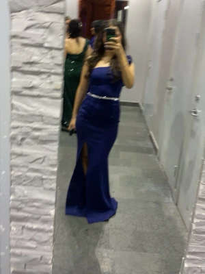 Marinblå långklänning med slits - Elegant marinblå långklänning med oneshoulder och volangdetalj på axeln. Klänningen har en markerad midja med pärlband och en hög slits framtill. Perfekt för bal eller festliga tillfällen. Materialet känns slätt och följsamt.