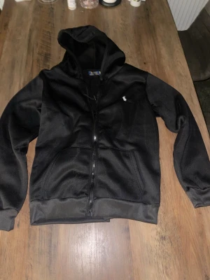 Polo Ralph Lauren - Snygg svart hoodie från Polo Ralph Lauren med dragkedja framtill och klassisk huva. Tröjan har fickor på magen och den ikoniska lilla loggan broderad på bröstet. Perfekt för en avslappnad och stilren look.