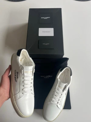 Saint Laurent vita sneakers - Snygga vita Saint Laurent sneakers i lågt snitt med svart detalj på hälen och klassisk logga på sidan. Skorna är tillverkade i slätt läder och har vita skosnören samt diskret guldtext på plösen. Perfekta för en clean och stilren look. Skickas samma dag som dem köps, skriv gärna vid frågor😊