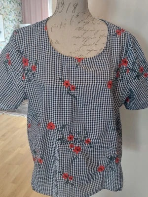 Rutig blus med broderade blommor - Supersöt blus i blåvitt rutigt tyg med broderade röda blommor. Blusen har rund halsringning och korta ärmar. Perfekt för dig som gillar vintagevibbar och detaljer. Materialet känns lätt och luftigt, bomull.