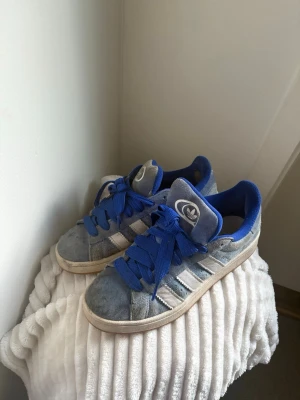 Blå Adidas Originals sneakers - Snygga Adidas Originals sneakers i blått och grått mocka med klassiska vita ränder på sidorna. Skorna har blå snören, blå insida och en vit sula. Perfekta för dig som gillar retrostil och vill ha något bekvämt till vardags.