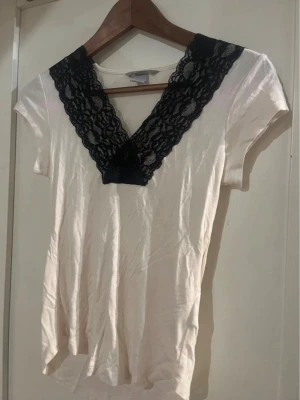 Vit topp med svart spetsdetalj - Vit kortärmad topp med djup v-ringning och bred svart spets längs halsen. Spetsen har ett blommigt mönster som ger en snygg kontrast mot den ljusa basen. Perfekt för dig som gillar detaljer och en feminin look.