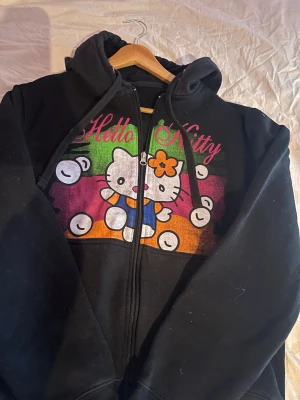 Hello Kitty zip - Svart hoodie med dragkedja och färgglatt Hello Kitty-tryck framtill. Motivet har rosa, grönt och orange bakgrund samt texten 'Hello Kitty' i rosa. Huva med snören och långa ärmar. Perfekt för dig som gillar gulliga och lekfulla plagg.