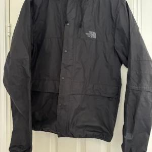 Svart vindjacka från The North Face - Säljer en svart vindjacka från The North Face med huva och dragkedja samt tryckknappar framtill. Jackan har logotyp på bröstet och ryggen, elastiska muddar och är tillverkad i lätt, vattenavvisande material. Perfekt för blåsiga dagar och enkel att packa med sig.