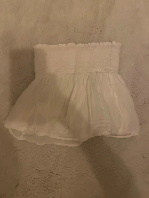 Vit axelbandslös bandeautopp - Söt vit bandeautopp med smockad överdel och volangig nederdel. Toppen är axelbandslös och har en luftig känsla, perfekt för varma dagar. Passar till både jeans och kjol för en trendig look. Strl 134