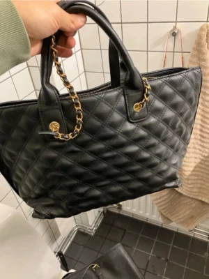 Svart quiltad handväska med guldkedja - Stilren svart handväska från NA-KD i quiltat konstläder med snygga guldfärgade kedjedetaljer. Väskan har dubbla handtag och dragkedja upptill. Perfekt för dig som gillar en klassisk och trendig look.