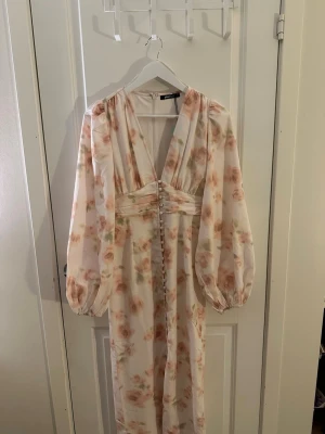 Beige blommig långklänning från Gina tricot  - Säljer en helt ny klänning från Gina tricot som är slutsåld i storlek 36. Säljer klänningen då den tyvärr var för stor för mig i storlek. Klänningen har djup v-ringning, puffiga långärmar och dekorativa knappar framtill. Perfekt klänning för midsommar☺️