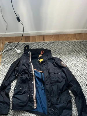 Svart vindjacka från Parajumpers - Snygg svart vindjacka från Parajumpers med flera praktiska fickor, dragkedja och tryckknappar. Jackan har en gul detalj vid halsen och ett emblem på ärmen. Insidan är blå med orange detaljer. Perfekt för blåsiga dagar och streetwear-stil. 