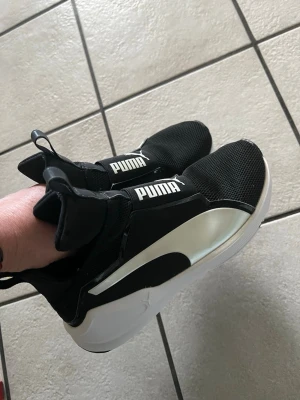 Svarta Puma sneakers med vit sula - Snygga svarta sneakers från Puma med vit sula och stor vit logga på ovansidan. Skorna har en sportig design med elastisk ovandel och tydlig Puma-logga på sidan. Ovandelen är i mesh och syntetmaterial för en modern look.