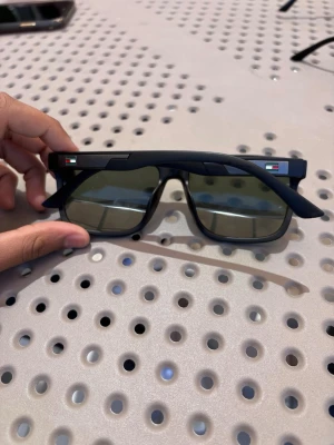 Svarta solglasögon från Tommyhilfiger  - Snygga svarta solglasögon med rektangulär form och mörka glas. På bågens sidor finns en liten flaggdetalj som ger en unik touch. Ramen är i plast och designen är modern och stilren, perfekt för soliga dagar.