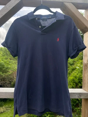 Marinblå pikétröja från Polo Ralph Lauren - Klassisk marinblå pikétröja från Polo Ralph Lauren med korta ärmar och röd broderad logga på bröstet. Använd så okej skick, storlek M 