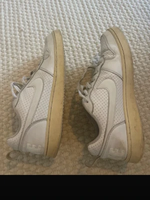 Vita Nike sneakers med perforerade detaljer - Fräscha vita Nike sneakers med perforerade paneler och klassisk swoosh på sidorna. Lågt skaft och beige sula ger en clean look. Perfekta för dig som gillar enkel och stilren design. Skorna har snörning och är tillverkade i syntetmaterial och textil.