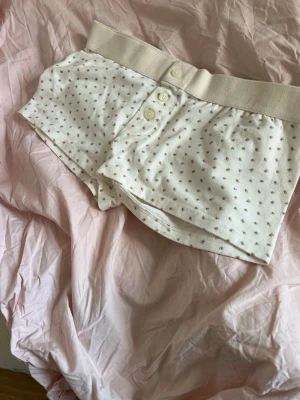 Vita kalsonger med små rosa stjärnor - Säljer ett par vita kalsonger med små rosa stjärnmönster och bred resår i midjan. Kalsongerna har tre knappar framtill och är gjorda i mjukt bomullstyg för extra komfort. Perfekta för dig som gillar en söt och enkel stil.