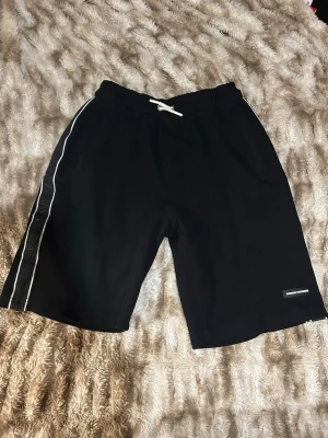 Svarta shorts från Armani Exchange - Svarta shorts från Armani Exchange med vit dragsko i midjan och en vit rand längs sidan. Materialet är mjukt och sportigt, perfekt för en avslappnad look. Diskret logga på vänster ben ger en clean vibe.