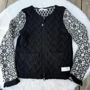 Svart kofta från Odd Molly (XS) - Säljer en svart kofta från Odd Molly med kabelstickad framsida och två fickor. Ärmarna är vita med svart blommönster och har ribbade muddar. Dragkedja framtill och dekorativt mönster i halsringningen. Perfekt för dig som gillar unika detaljer.