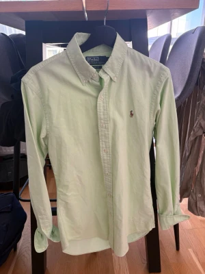 Ljusgrön skjorta från Polo Ralph Lauren - Klassisk ljusgrön skjorta från Polo Ralph Lauren med button-down krage och broderad logga på bröstet. Skjortan är långärmad och har knappar framtill. Tillverkad i mjuk bomull för en bekväm känsla.