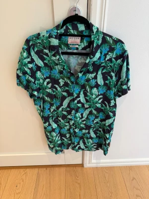 Guess Skjorta – Regular Fit, Strl S – Kanonskick - Snygg och trendig skjorta från Guess i mörkblått med ett stilfullt tropiskt mönster av gröna blad. En perfekt skjorta för sommarens fester, semestern eller för dig som vill sticka ut med en avslappnad men välklädd look.  ✔ Märke: Guess ✔ Storlek: S ✔ Passform: Regular Fit ✔ Färg: Mörkblå med grönt tropiskt bladmönster ✔ Kanonskick  Skjortan är väl omhändertagen och kommer från ett djur- och rökfritt hem.