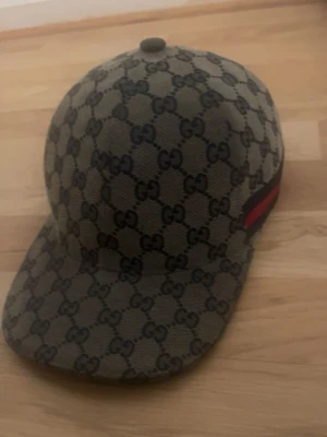 Gucci keps med GG-mönster - Säljer en snygg keps från Gucci med klassiskt GG-monogram i beige och mörkbrunt. Kepsen har en böjd skärm och en ikonisk röd-grön rand på sidan. Tillverkad i canvasmaterial med mörkblått innerfoder och tydlig Gucci-label på insidan.