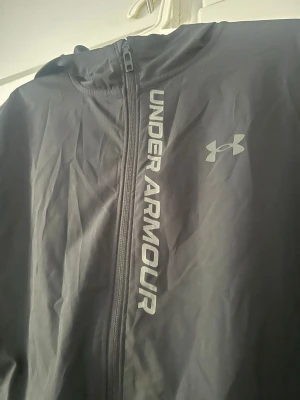 Svart vindjacka från Under Armour storlek s - Snygg svart vindjacka från Under Armour med huva och hel dragkedja. Jackan har logotyp och text på framsidan samt diskreta reflexdetaljer på ryggen. Materialet är lätt och vindtätt, perfekt för aktiva dagar utomhus.