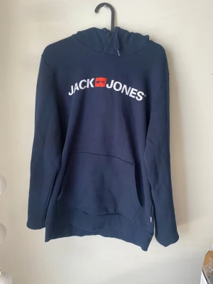Mörkblå hoodie från Jack & Jones - Snygg mörkblå hoodie från Jack & Jones med stor vit logga och röd detalj på bröstet. Klassisk känguruficka framtill och huva. Tillverkad i mjukt bomullsmaterial, perfekt för en avslappnad stil.
