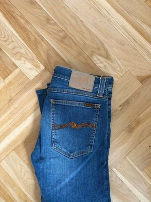 Blå jeans från Nudie Jeans - Snygga blå jeans från Nudie Jeans med klassisk femficksdesign och orangea sömmar på bakfickan. Jeansen har en rak passform och är tillverkade i denim av bomull. Perfekta för en avslappnad och stilren look.