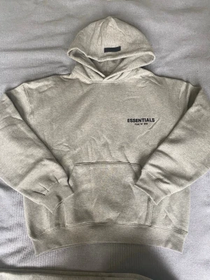 Grå Essentials Fear of God hoodie - Säljer en grå hoodie från Essentials Fear of God med svart logga på bröstet och stor text på ryggen. Hoodien har huva, magficka och ribbade muddar vid ärmar och nederkant. Tillverkad i mjukt bomullsmaterial, perfekt för en avslappnad streetwear-look.