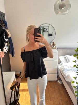 Svart offshoulder blus med volang - Säljer en svart offshoulder blus med bred volang upptill och tunna axelband. Blusen har en luftig och flowy passform, perfekt för varma dagar. Enkel och stilren design som passar till mycket.