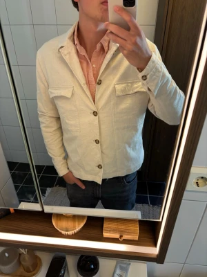 Ljusbeige kavaj från Selected Homme - Stilren ljusbeige kavaj från Selected Homme med klassisk krage, bröstfickor med lock och knäppning framtill. Tillverkad i ett lätt tyg som passar perfekt till jeans eller chinos. Enkel och modern design som ger en avslappnad men snygg look.