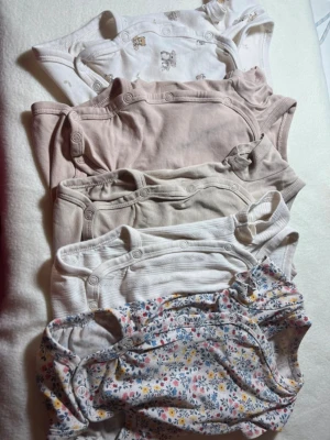 5-pack neutrala och blommiga bodys - Säljer ett 5-pack bodys i mjuk bomull. Färgerna är beige, vitt och ljusgrått, samt en med blommigt mönster i rosa, blått och gult. Några har tryckknappar framtill och en har sött djurmönster. Perfekta för en enkel och söt vardagslook.