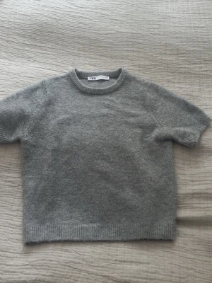 Grå stickad t-shirt från Zara - Kortärmad stickad t-shirt i mjuk grå ullmix från Zara. Tröjan har rund halsringning och ribbade kanter vid ärmslut och nederkant. Enkel och stilren design som passar perfekt till lager på lager.