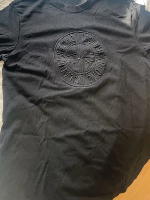 Svart Stone Island t-shirt med logga - Snygg svart t-shirt från Stone Island med stor, diskret broderad logga på bröstet. Klassisk rund hals och korta ärmar. Tillverkad i mjuk bomull som känns skön mot huden. Perfekt för dig som gillar stilrena plagg med exklusiv känsla.
