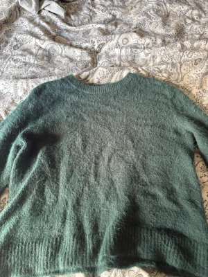 Grön fluffig stickad tröja - Mysig stickad tröja i en djup grön färg med fluffig yta. Tröjan har rund halsringning, långa ärmar och ribbstickade muddar vid ärmslut och nederkant. Perfekt för kalla dagar när du vill vara både bekväm och snygg.