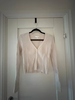 Beige ribbad kofta från H&M - Snygg beige ribbad kofta från H&M med v-ringning och knäppning framtill. Modellen är croppad med långa ärmar och smal passform, perfekt att styla över ett linne eller t-shirt. Materialet är mjukt och stretchigt för extra komfort.