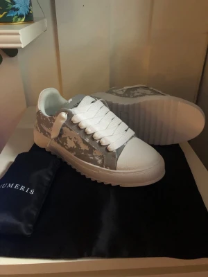 Numeris vita sneakers med camomönster - Snygga vita sneakers från Numeris med grått camomönster på sidorna och kraftig räfflad sula. Skorna har vita skosnören och detaljer i textil och syntet. Perfekta för dig som vill sticka ut med en unik look.