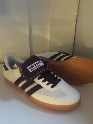 Adidas Samba OG vita/bruna sneakers - Klassiska Adidas Samba OG sneakers i vitt och mörkbrunt med ikoniska tre ränder på sidan. Ovandelen är i läder och mocka, med gummisula i brunt. Tungan har Adidas-logga och modellen har retrovibbar. Perfekta för dig som gillar streetwear och tidlös stil.